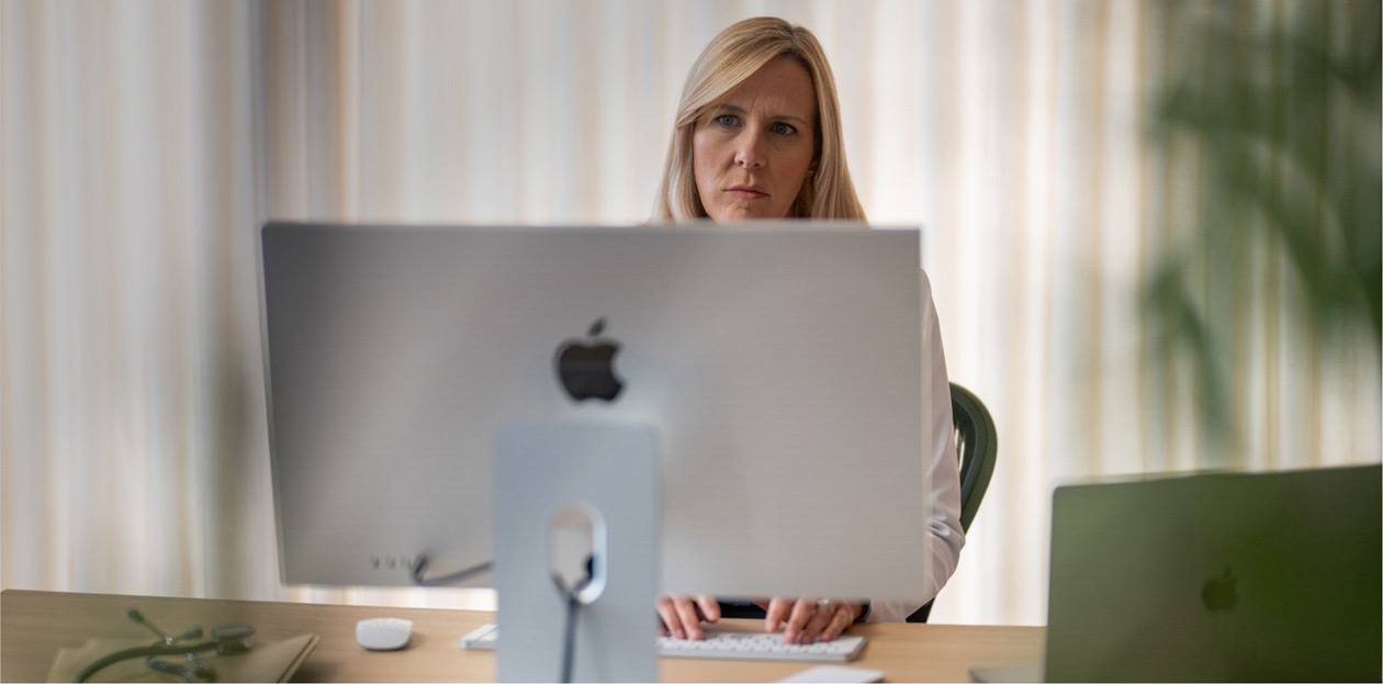 Eine Mitarbeiterin eines Gesundheitsdienstleisters schaut hinter ihrem Schreibtisch auf einen iMac Bildschirm.