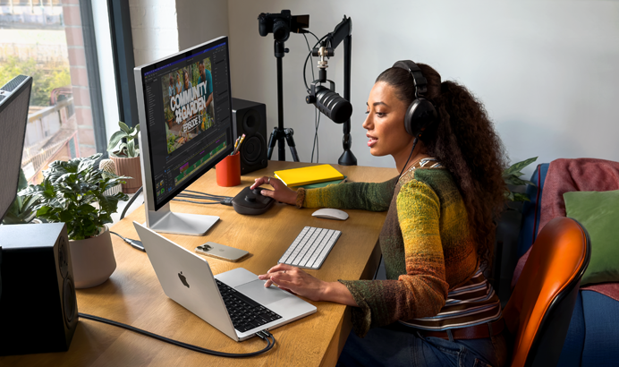 Moteris namuose dirba prie stalo naudodama „MacBook Pro“, kuris prijungtas prie dviejų išorinių ekranų, dėvi ausines ir kalba į mikrofoną