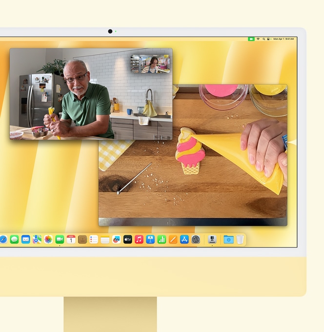 Жовтий iMac. Демонстрація функції «Вид на стіл». На екрані показано кондитерку на відеовиклику з іншою людиною. На екрані видно обличчя кондитерки та чоловіка. На іншому екрані показано руки пекаря, який прикрашає печиво глазур’ю