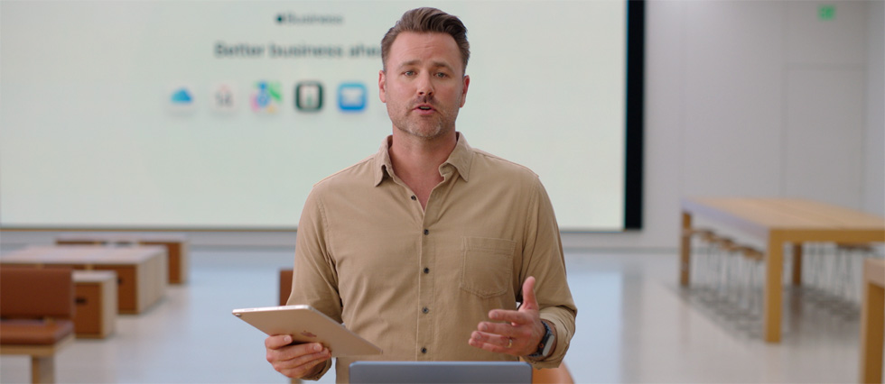 發表人手持 iPad 站在放著 MacBook 的桌前發言，同時大螢幕顯示著一系列商務相關的 app 圖像。
