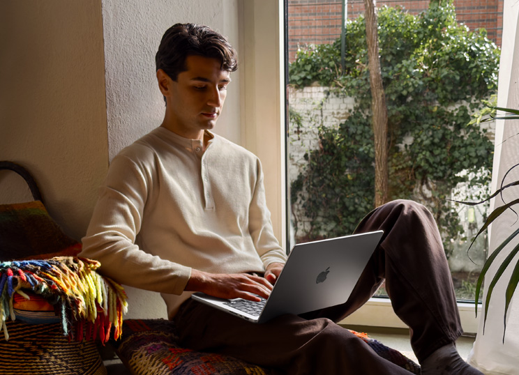 Iemand die met een MacBook naast een raam op de vloer zit.