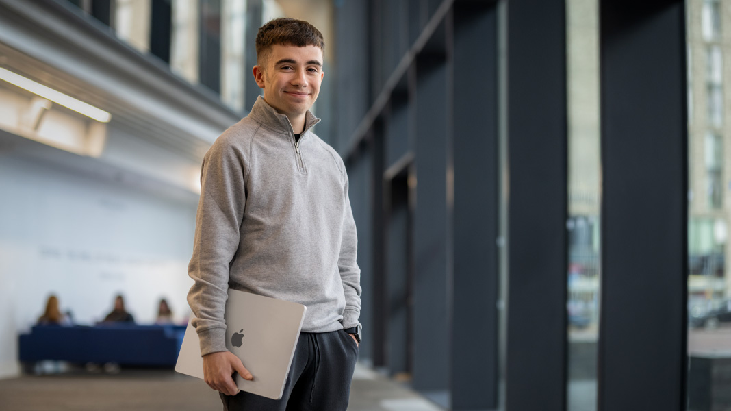 Ein Student steht mit einem MacBook in der Hand im Korridor eines modernen Gebäudes und lächelt in die Kamera