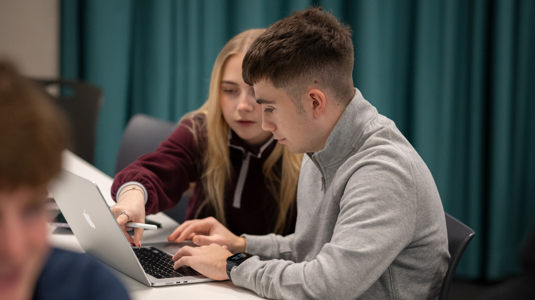 Ein Schüler und eine Schülerin arbeiten auf einem MacBook zusammen, sie zeigt auf den Bildschirm, während er tippt