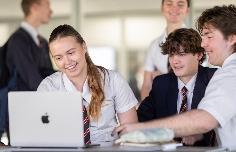 Eine Gruppe von Schüler:innen in Schuluniformen versammelt sich um ein MacBook, lächelt und arbeitet zusammen.