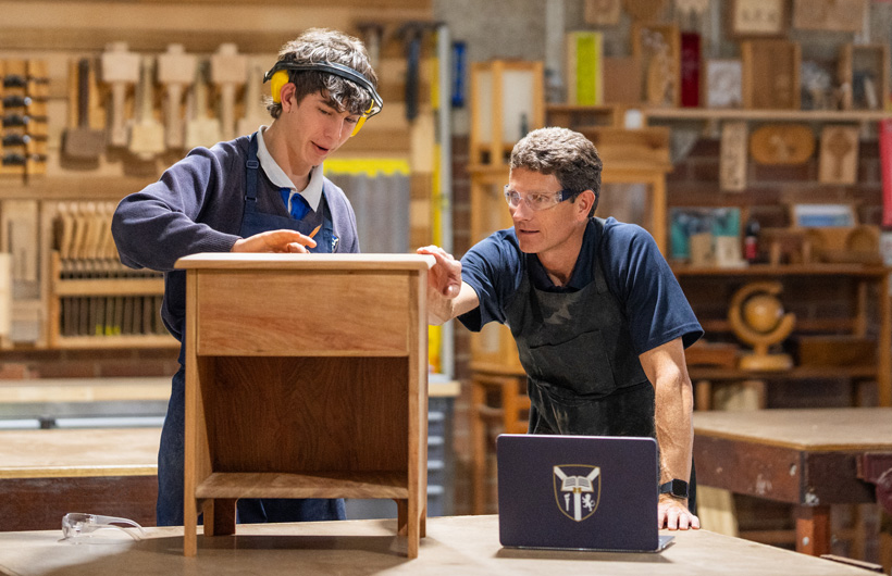 Ein Holzwerklehrer und ein Schüler begutachten einen kleinen Holzschrank. Ein schuleigener Laptop mit Logo der Schule liegt auf der Werkbank vor ihnen.