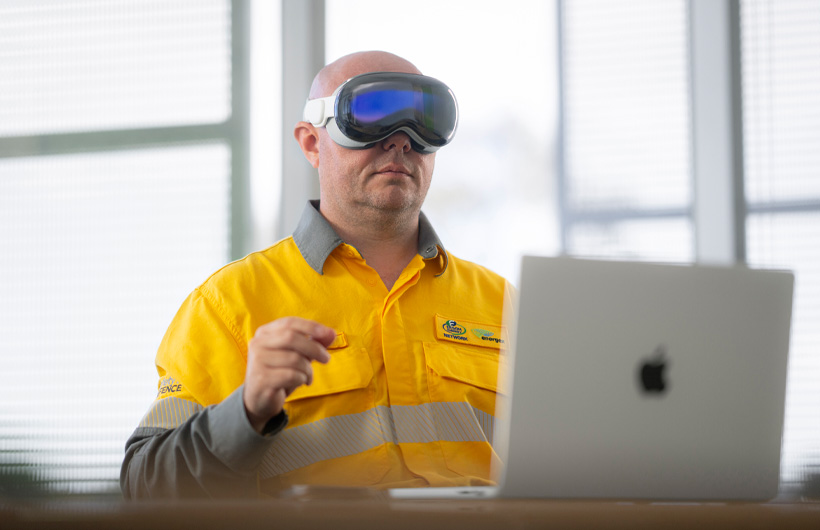 An employee wears Apple&nbsp;Vision&nbsp;Pro and makes a pinching gesture with their hands, while a MacBook&nbsp;Pro in Silver sits open on the desk