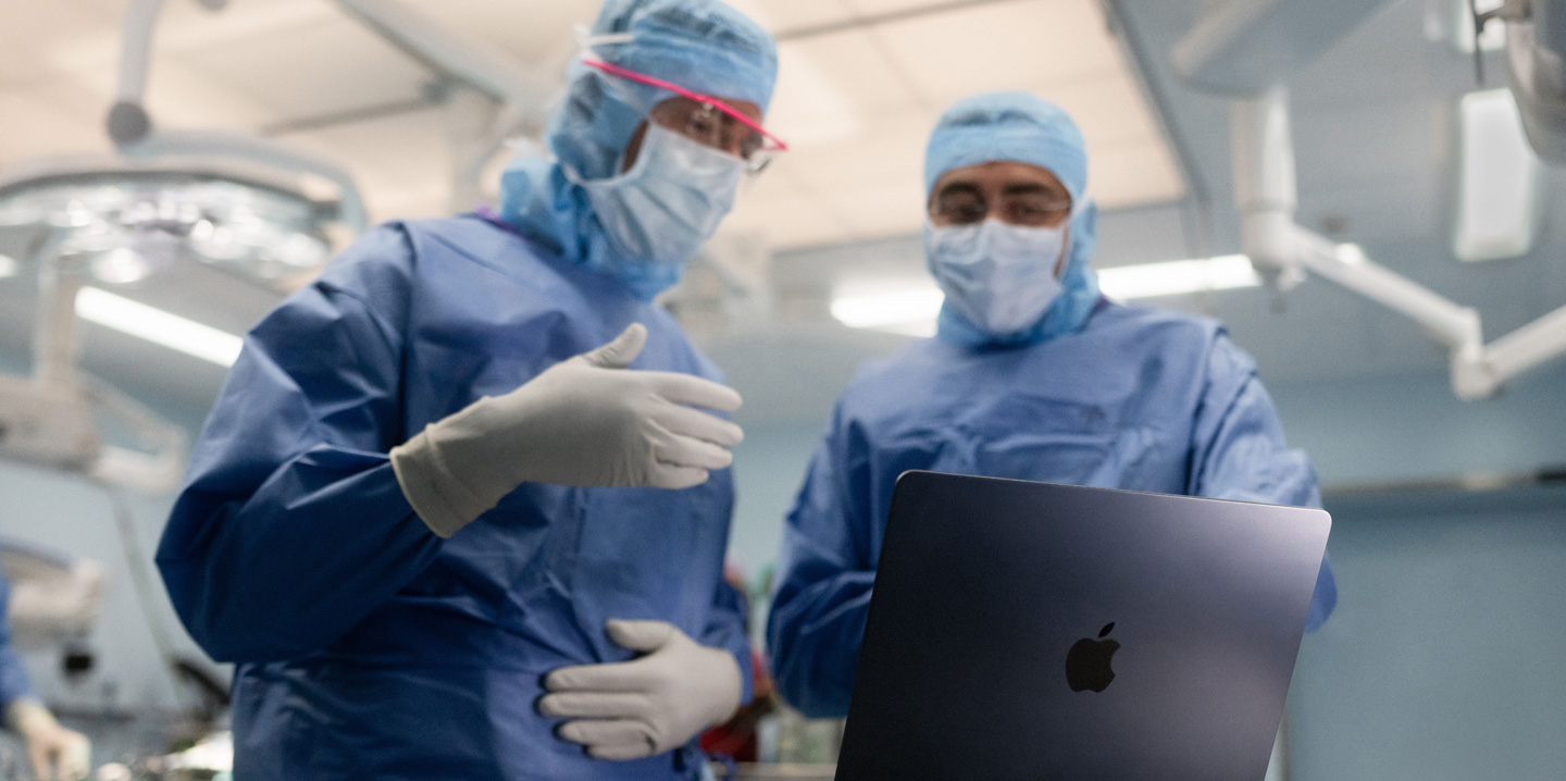 Zwei Chirurgen im Operationssaal schauen auf ein MacBook, wobei einer eine Geste mit der Hand macht, während der andere beobachtet