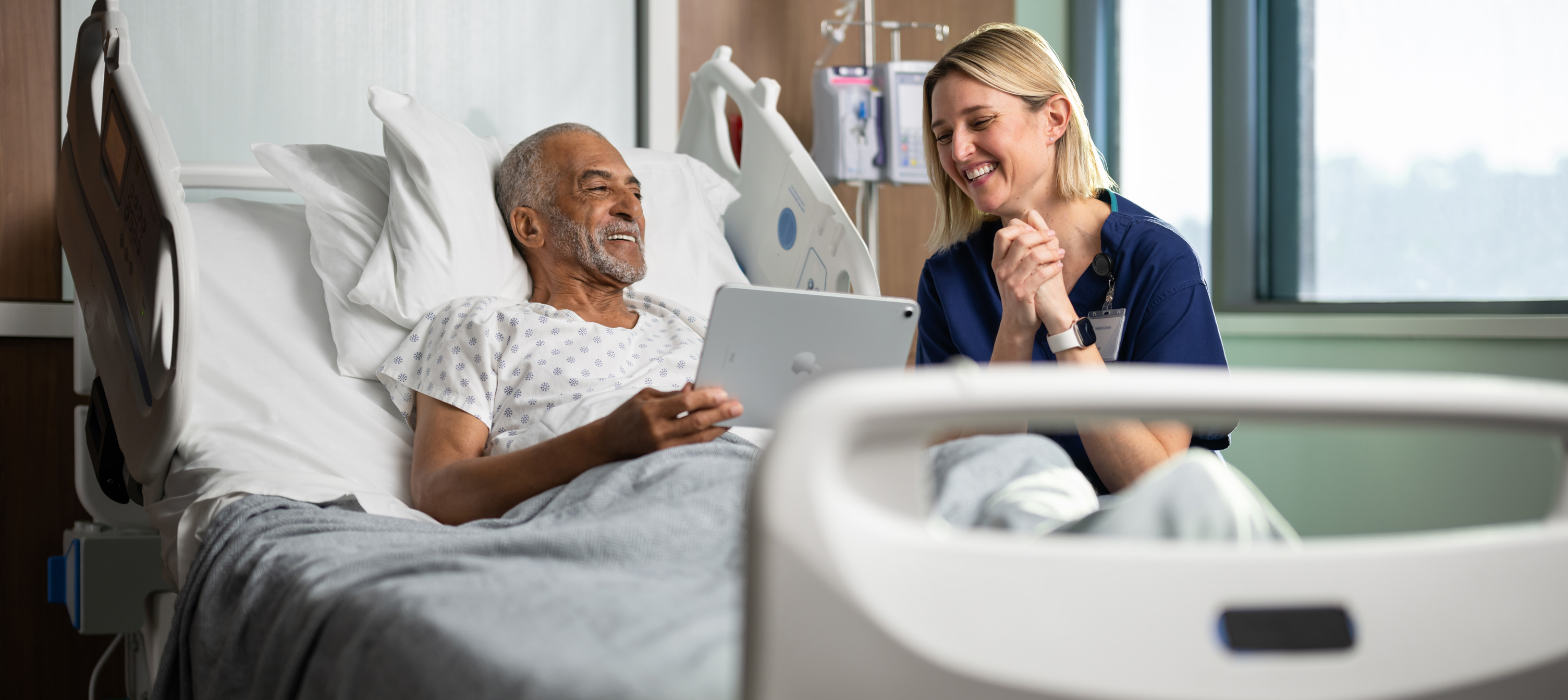 En manlig patient som sitter i en sjukhussäng. Han ler medan han tittar på en iPad tillsammans med en kvinnlig sjuksköterska.